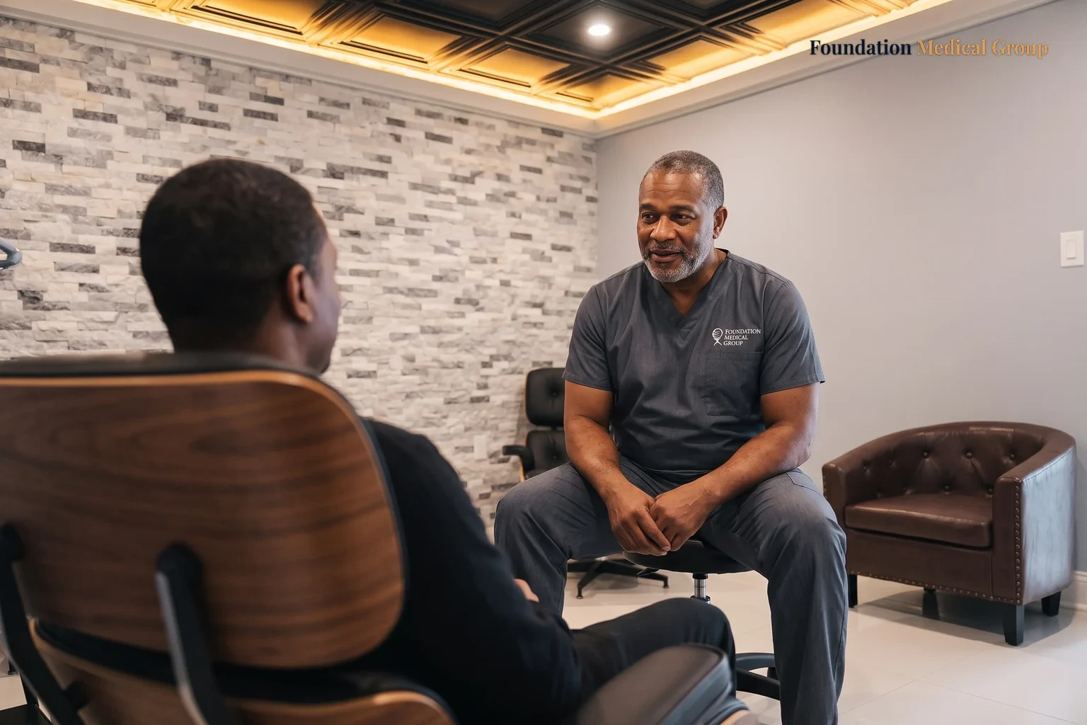 A clinician in charcoal scrubs sitting forward in a treatment room, talking with a patient who is seen from behind in a walnut lounge chair