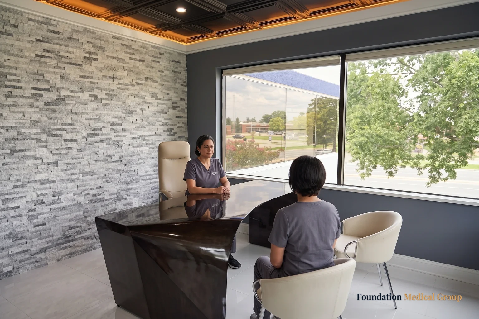 A wide view of a corner consult office where a clinician sits behind a dark desk and a patient listens from a cream chair by the window