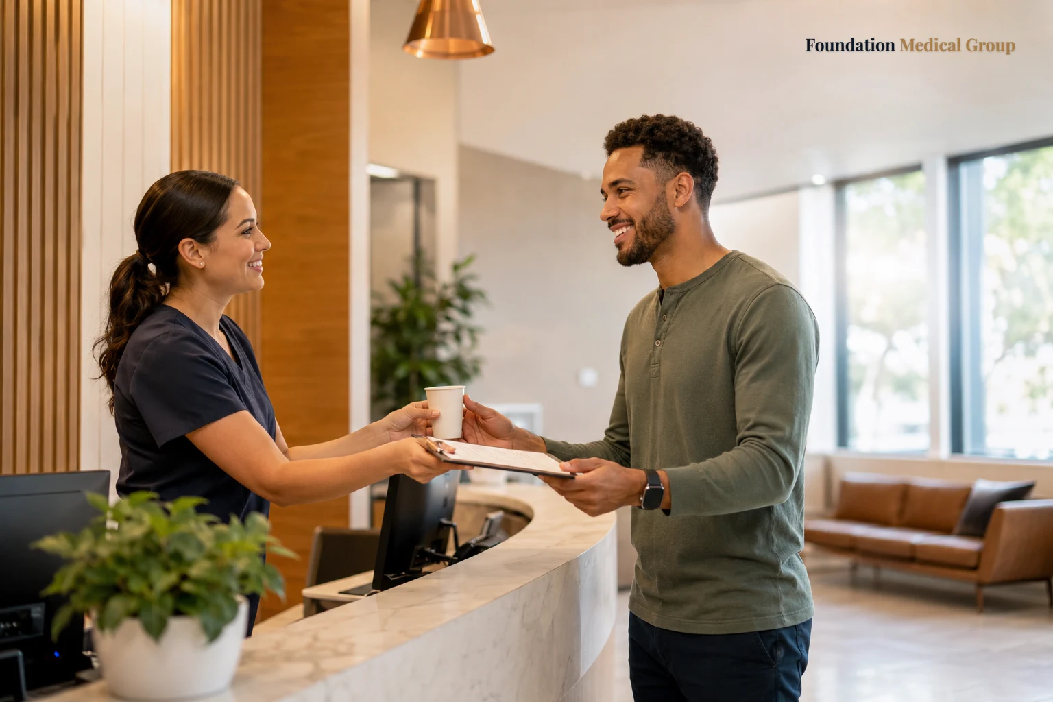 A friendly front-desk staff member hands a smiling young man a clipboard and a paper cup of water at a bright, tidy chec