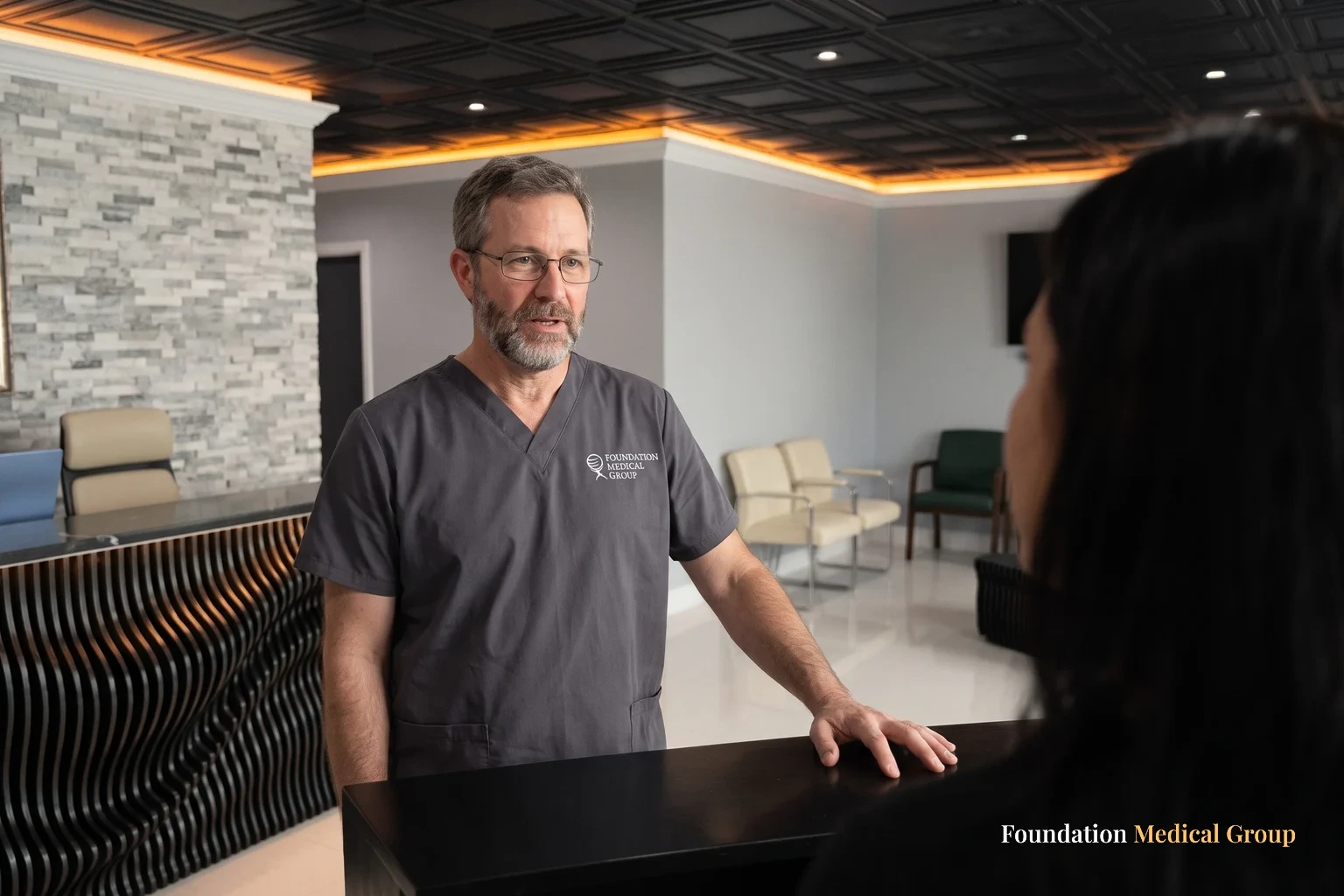A clinician standing behind the ribbed reception desk, speaking with a patient whose shoulder fills the foreground