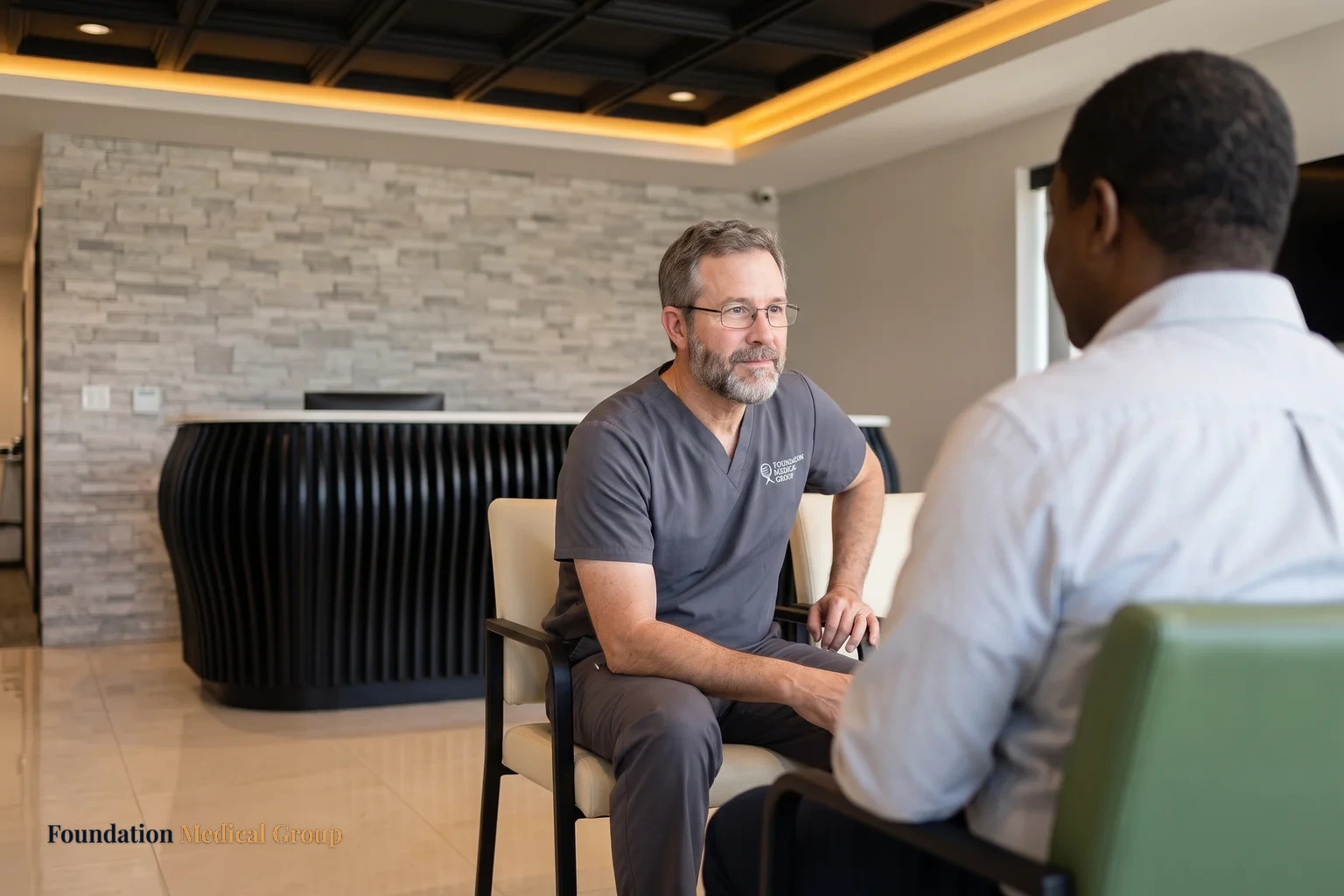 A clinician seated in the reception area, leaning forward at eye level with a patient seen from behind