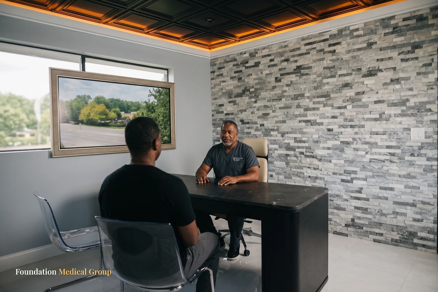 A wide consult office where a clinician sits behind a dark stone desk and a patient faces him from a clear chair