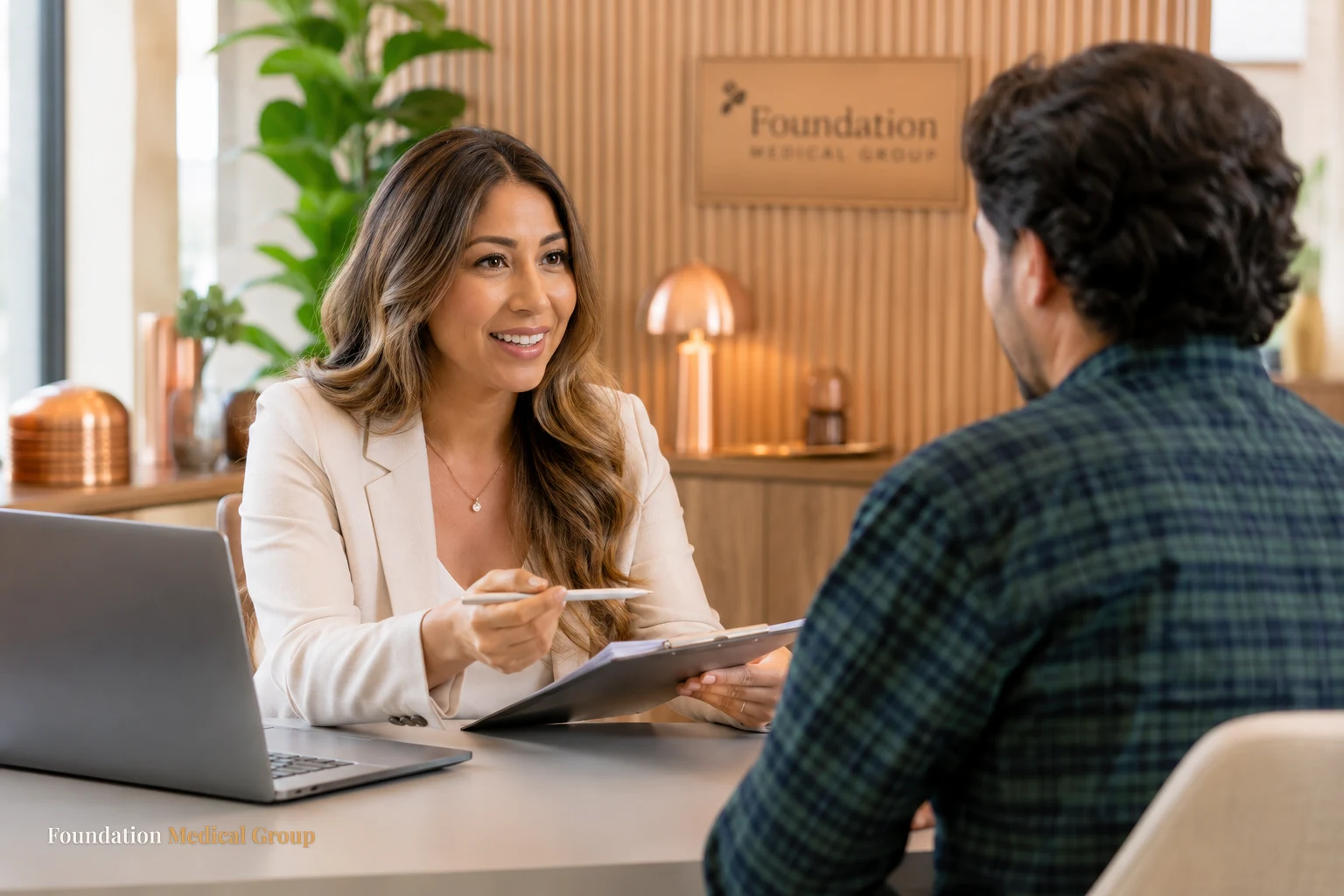 A caring clinic coordinator explains coverage options warmly to a patient across a bright desk with a laptop and copper-
