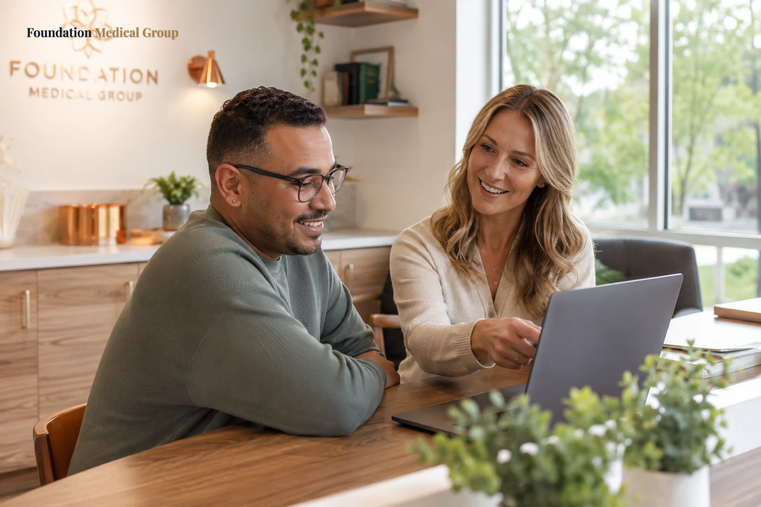 A relieved patient smiles as a helpful staff member points encouragingly at a laptop while explaining coverage in a sunn